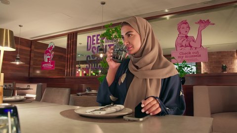 احتساء المشروب المفضل، ايماءات السعادة و السرور ,اجراء مكالمة هاتفية، استخدام جهاز تقني حديث ومتطور، امراه عربيه خليجيه اماراتية ترتدي العباءة و الحجاب تجلس فى احد المقاهي تمسك الهاتف المحمول وكوب من العصير، العمل عن بعد، التواصل وتبادل الأحاديث مع الأهل والأصدقاء