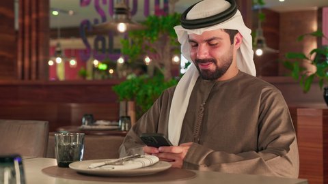 Exchanging conversations, communication, and news with family and friends, browsing applications and social media platforms, working remotely, an Arab Gulf Emirati man wearing a kandura and a white ghutrah sits in a cafe holding a mobile phone, using a modern and advanced technological device.