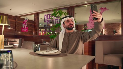 استخدام الأجهزة والتقنيات الحديثة، التقاط صور سيلفي تذكارية عبر الجوال, شاب عربي خليجي اماراتي يرتدي الكندورة و الغتره يجلس فى احد المقاهي يقوم بحمل الهاتف المحمول, اجراء مكالمة فيديو عبر تطبيقات ومواقع السوشال ميديا, قضاء وقت ممتع فى الخارج