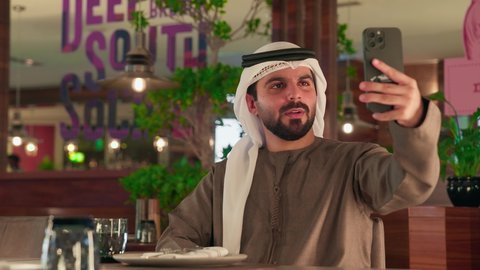 التقاط صور سيلفي تذكارية عبر الجوال، استخدام الأجهزة والتقنيات الحديثة, شاب عربي خليجي اماراتي يرتدي الكندورة و الغتره يجلس فى احد المقاهي يقوم بحمل الهاتف المحمول, اجراء مكالمة فيديو عبر تطبيقات ومواقع السوشال ميديا, قضاء وقت ممتع فى الخارج