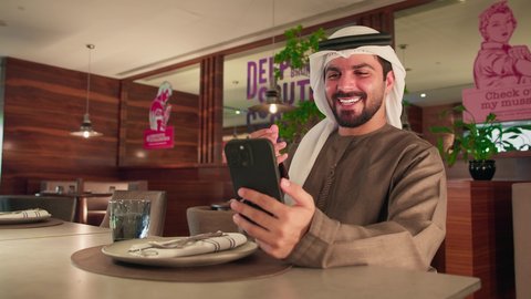 مفهوم القبول و الموافقة ، اجراء مكالمة فيديو, الاطمئنان على الاهل والاصدقاء ، تصفح مواقع التواصل الاجتماعي , رجل عربي خليجي اماراتي يرتدي الكندورة و الغتره يجلس فى احد المقاهي  يقوم باستخدام الهاتف المحمول, دمج التقنيات الحديثة فى الحياة اليومية, ايماءات السعادة و السرور