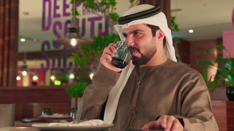 ايماءات السعادة و السرور ، التواصل وتبادل الأحاديث مع الأهل والأصدقاء,اجراء مكالمة هاتفية، استخدام جهاز تقني حديث ومتطور، احتساء المشروب المفضل، رجل عربي خليجي اماراتي يرتدي الكندورة والغترة البيضاء يمسك بيده الهاتف المحمول وكوب من العصير، العمل عن بعد