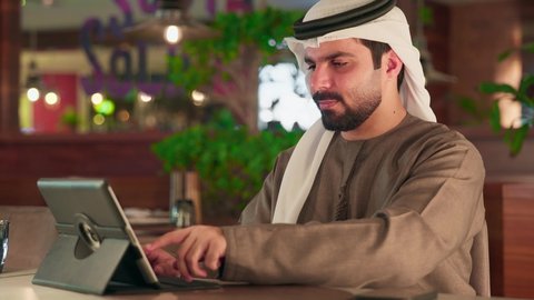 He uses the tablet device to accomplish projects and tasks. An Emirati Gulf Arab man wearing a kandura and ghutrah sits in a cafe using a portable tablet, following social media, making video calls, and live streaming online, enjoying his time.