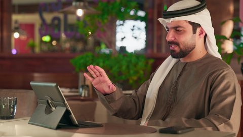إجراء مكالمة فيديو، رفع الكف للأعلى بايماءات التحية والسلام، المتابعة والإشراف على العمل عبر جهاز التابلت, ايماءات السرور والسعادة, العمل عن بعد من خلال الانترنت, رجل عربي خليجي اماراتي يرتدي الكندورة و الغتره يجلس في مقهى
