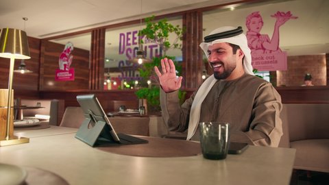 المتابعة والإشراف على العمل عبر جهاز التابلت, رفع الكف للأعلى بايماءات التحية والسلام, إجراء مكالمة فيديو, ايماءات السرور والسعادة, العمل عن بعد من خلال الانترنت, رجل عربي خليجي اماراتي يرتدي الكندورة و الغتره يجلس في مقهى