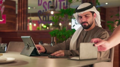 رجل عربى خليجي اماراتي يرتدي الكندورة و الغترة  يقوم بدفع الحساب عبر الهاتف المحمول، العمل على الجهاز اللوحي، استخدام الوسيلة التقنية الحديثة في الدفع الالكتروني، سهولة الدفع الالي في الكافيهات الاماراتية