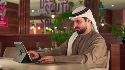 العمل على الجهاز اللوحي، استخدام الوسيلة التقنية الحديثة في الدفع الالكتروني، سهولة الدفع الالي في الكافيهات الاماراتية, رجل عربى خليجي اماراتي يرتدي الكندورة و الغترة  يقوم بدفع الحساب عبر الهاتف المحمول،