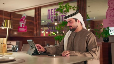 سهولة الدفع الالي في الكافيهات الاماراتية, رجل عربى خليجي اماراتي يرتدي الكندورة و الغترة  يقوم بدفع الحساب عبر الهاتف المحمول, استخدام الوسيلة التقنية الحديثة في الدفع الالكتروني، العمل على الجهاز اللوحي