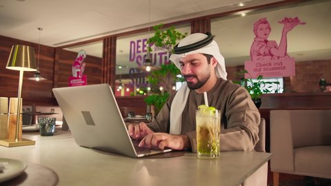 Clicking on the keyboard, creating textual content, integrating modern technology into practical life, remote work, typing on a laptop, the concept of data entry, a photo of an Emirati Gulf Arab man wearing a kandura and ghutrah sitting in a cafe using a laptop, enjoying drinks.