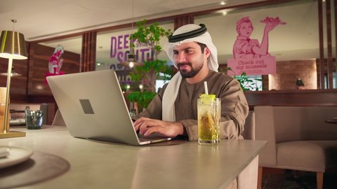 صناعة المحتويات النصية، دمج التكنولوجيا الحديثة بالحياة العملية، العمل عن بعد، الكتابة على جهازاللابتوب، مفهوم ادخال البيانات، النقر على لوحة المفاتيح، تصوير لرجل عربي خليجي اماراتي يرتدي الكندورة و الغترة يجلس فى احدي الكافيهات يستخدم الحاسوب المحمول، الاستمتاع بتناول المشروبات