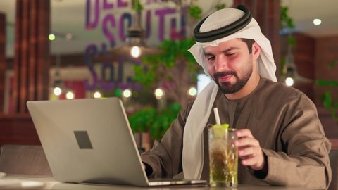 دمج التكنولوجيا الحديثة بالحياة العملية، العمل عن بعد، الكتابة على جهازاللابتوب، مفهوم ادخال البيانات، النقر على لوحة المفاتيح، صناعة المحتويات النصية، تصوير مقرب لرجل عربي خليجي اماراتي يرتدي الكندورة و الغترة يجلس فى احدي الكافيهات يستخدم  الحاسوب المحمول، الاستمتاع بتناول المشروبات