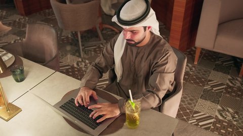 Remote work, typing on a laptop, the concept of data entry, clicking on the keyboard, creating textual content, an Arab Gulf Emirati man wearing a kandura and ghutrah sitting in a cafe using a laptop, integrating modern technology into practical life, enjoying beverages.