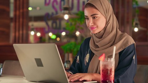 The use of modern and advanced devices, remote business management, a close-up shot of an Emirati Gulf Arab woman wearing an abaya and hijab sitting in a cafe using a laptop, integrating technology with work, a Saudi woman completing the required tasks via laptop, enjoying refreshing drinks.