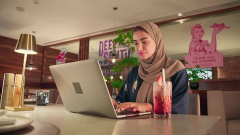 ادارة الأعمال عن بعد، استخدام الأجهزة الحديثة والمتطورة، امراة عربية خليجية اماراتية ترتدي العباءة والحجاب تجلس فى احدي الكافيهات تقوم باستخدام الحاسوب المحمول، دمج التكنولوجيا بالعمل، سيده سعودية تقوم بإنجاز المهام المطلوبة عبر اللابتوب، الاستمتاع بتناول المشروبات المنعشة