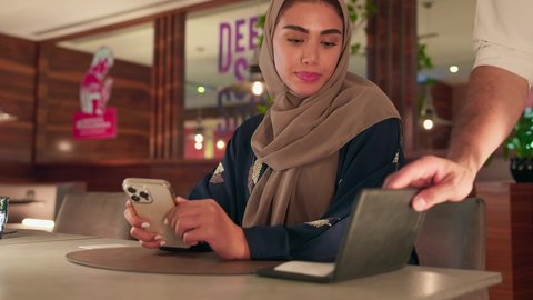 احدي الكافيهات الحديثة في المملكة العربية السعودية، أسلوب الضيافة الراقية في المقاهي السعودية، سيده سعودية امارتية تقوم بالدفع الالكتروني عبر بطاقة الائتمان ،تصوير مقرب  لنادل يقدم الماكينة لامرأة سعودية ترتدي العباءة والحجاب وهي تستخدم هاتفها للدفع