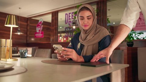 سيده سعودية امارتية تقوم بالدفع الالكتروني عبر بطاقة الائتمان ، أسلوب الضيافة الراقية في المقاهي السعودية ،تصوير مقرب  لنادل يقدم الماكينة لامرأة سعودية ترتدي العباءة والحجاب وهي تستخدم هاتفها للدفع ، احدي الكافيهات الحديثة في السعودية
