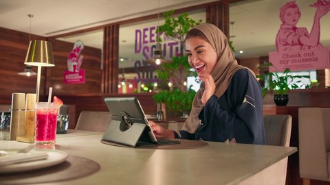 ايماءات السعادة و السرور، الدردشة مع الاهل و الاصدقاء، دمج التقنية في الحياة اليومية والعملية، امرأة سعودية ترتدي العباءة والحجاب تجلس فى المقهي تقوم باجراء مكالمة فيديو عبر جهاز التابلت، امرأة عربية خليجية اماراتية تقوم باستخدام الاجهزة التقنية الحديثة ، تصفح مواقع ووسائل التواصل الاجتماعي