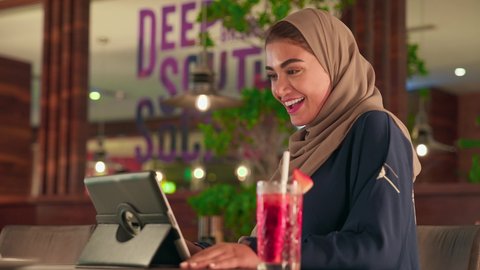 Chatting with family and friends, integrating technology into daily and professional life, a Saudi woman wearing an abaya and hijab sitting in a cafe making a video call via a tablet, an Emirati Gulf Arab woman using modern technological devices, browsing social media sites, enjoying refreshing drinks.