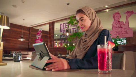 دمج التقنية في الحياة اليومية والعملية، تصوير لامرأة عربية خليجية سعودية ترتدي العباءة والحجاب تجلس فى الكافية تقوم بانجاز مهام العمل عبر جهاز التابلت، امرأة عربية خليجية اماراتية تقوم باستخدام الاجهزة التقنية الحديثة ، تصفح مواقع ووسائل التواصل الاجتماعي، مفهوم انجار المشاريع عن بعد ، الاستمتاع بتناول المشروبات المنعشة