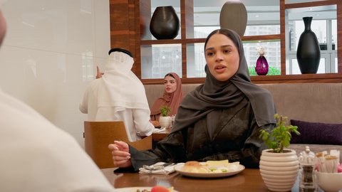 تواصل وحوار بين أفراد الأسرة على المائدة، الاستمتاع بتناول وجبة الافطار ، قضاء وقت ممتع في الخارج, امراة عربية خليجيه اماراتية محجبه ترتدي العباءة تجلس برفقة زوجها في احدى المطاعم الاماراتية، ايماءات السعادة و السرور
