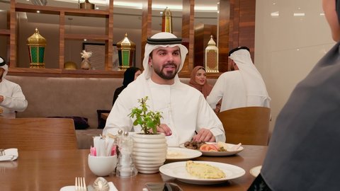 Family dialogues and discussions, enjoying breakfast, spending enjoyable time outdoors, the concept of intimacy and love among family members, an Emirati Gulf Arab man wearing a kandura and ghutrah sitting with his wife in one of the Emirati restaurants, gestures of happiness and joy.