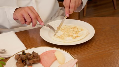 Spending a good time in the restaurant, a close-up shot of an Emirati Gulf Arab man wearing a kandura enjoying a meal of eggs, holding a knife and fork while eating, with a calm and comfortable breakfast atmosphere.
