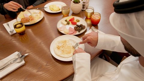 الاستمتاع بالغداء في الخارج, قضاء أوقات ممتعة في المطعم, مطاعم و كافيهات الامارات العربية,  تصوير مقرب لشاب عربي خليجي اماراتي الكندورة و الغتره يجلس على المائدة برفقة زوجته يستمتع بتناول وجبة البيض يمسك بيدة السكين والشوكة اثناء تناول الطعام