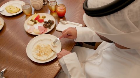 A calm and relaxing breakfast atmosphere, a close-up shot of an Emirati Gulf Arab man wearing a kandura enjoying a meal of eggs, holding a knife and fork while eating, having a good time in the restaurant.