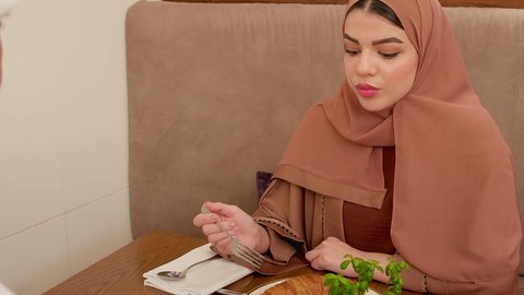 الاستمتاع بوجبة الإفطار في أجواء المطعم المريحة، تبادل الأحاديث والاخبار, تصوير مقرب لامراة عربيه خليجيه اماراتية ترتدي الحجاب و العباءة تجلس على المائدة في إحدى المطاعم الراقية تقوم بتناول الطعام  و التحدث مع شخص ما