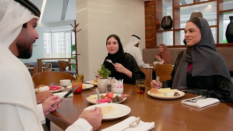 A lively family gathering and friendly discussions, a table full of food and drinks, gestures of happiness and joy, a weekend, enjoying drinks, an Emirati Gulf Arab family dressed in traditional attire sitting in a restaurant having breakfast.