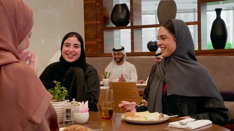 مناقشات أسرية ودية على المائدة، سفرة مليئة بالمأكولات والمشروبات، ايماءات السعادة والسرور، عطلة نهاية الاسبوع، قضاء وقت ممتع، عائله عربيه خليجيه اماراتية ترتدي الحجاب و العباءة يجلسون فى احد المطاعم يتناولون الافطار