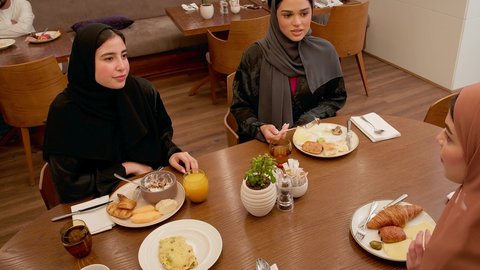 تفاعل الأسرة وتبادل الأحاديث أثناء الأكل، سفرة مليئة بالمأكولات والمشروبات، ايماءات السعادة والسرور، عطلة نهاية الاسبوع، قضاء وقت ممتع، عائله عربيه خليجيه اماراتية ترتدي الحجاب و العباءة يجلسون فى احد المطاعم يتناولون الافطار