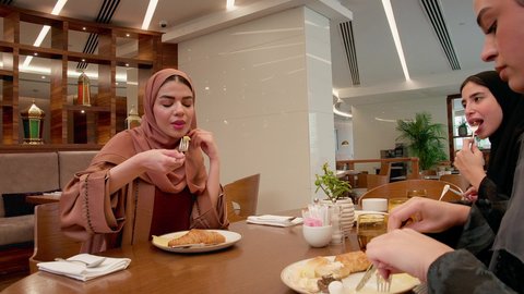A table full of food and drinks, a family atmosphere filled with love, gestures of happiness and joy, the weekend, spending enjoyable time, an Emirati Gulf Arab family wearing hijabs and abayas sitting in a restaurant having breakfast.
