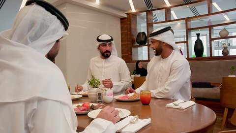 The exchange of conversation among family members during a meal, gestures of happiness and joy, the weekend, a happy family atmosphere, having a good time, an Emirati Gulf Arab family wearing the kandura and ghutrah sitting in a restaurant having breakfast.