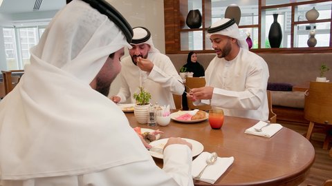 Weekend, happy family atmosphere, a dining table filled with the most delicious foods and drinks, having a good time, an Emirati Gulf Arab family wearing kandura and ghutrah sitting in a restaurant having breakfast, gestures of happiness and joy.