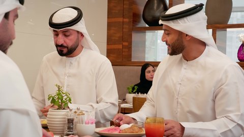 Happy family atmosphere, a dining table filled with the most delicious foods and drinks, having a good time, a close-up shot of an Emirati Gulf Arab family wearing kandura and ghutrah sitting in a restaurant having breakfast, gestures of happiness and joy.