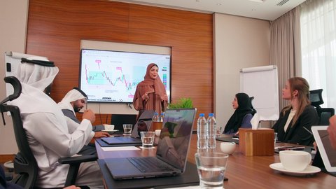 تحقيق أهداف الشركة، موظفة اماراتية تعرض البيانات الاحصائية على الشاشة، ضمان فهم واضح وشامل للأعمال، استخدام الأجهزة التقنية الحديثة في المكاتب, الاتفاق على المشاريع التجارية، اجتماع عمل بين موظفين عرب خليجيين اماراتيين مع رجال أعمال أجانب في غرفة الاجتماعات