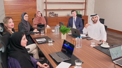 شرح وتوضيح العمليات والمشاريع والصفقات التجارية، التفاهم والتعاون بين الزملاء لتحقيق أهداف الشركة, نقاشات خاصة بالعمل, استخدام الحاسوب المحمول، اجتماع عمل مع موظفين عرب خليجيين اماراتيين مع رجال أعمال أجانب في غرفة الاجتماعات، ضمان فهم واضح وشامل للأعمال