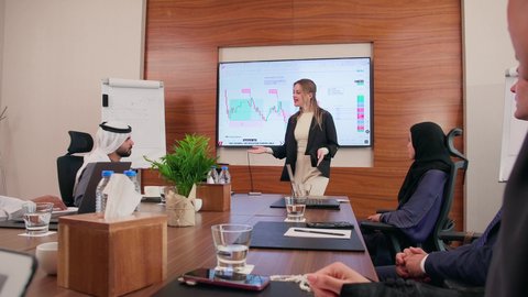 شرح وتوضيح العمليات والمشاريع التجارية، تحقيق أهداف الشركة، موظفة اجنبية تعرض البيانات الاحصائية على الشاشة، ضمان فهم واضح وشامل للأعمال، استخدام الأجهزة التقنية الحديثة في المكاتب, اجتماع عمل بين موظفين عرب خليجيين اماراتيين مع رجال أعمال أجانب في غرفة الاجتماعات