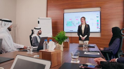 توضيح العمليات والمشاريع التجارية، تحقيق أهداف الشركة، موظفة اجنبية تعرض البيانات الاحصائية على الشاشة، ضمان فهم واضح وشامل للأعمال، استخدام الأجهزة التقنية الحديثة في المكاتب, اجتماع عمل بين موظفين عرب خليجيين اماراتيين مع رجال أعمال أجانب في غرفة الاجتماعات