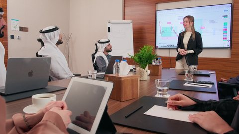 شرح وتوضيح العمليات والمشاريع التجارية، تحقيق أهداف الشركة، موظفة اجنبية تعرض البيانات الاحصائية على الشاشة، ضمان فهم واضح وشامل للأعمال، استخدام الأجهزة التقنية الحديثة في المكاتب, اجتماع عمل بين موظفين عرب خليجيين اماراتيين مع رجال أعمال أجانب في غرفة الاجتماعات