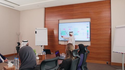 The use of modern technological devices in offices, a comfortable work environment in Emirati companies, an Emirati employee presenting statistical data on the screen, agreement on business projects, a business meeting between Arab Gulf Emirati employees and foreign businessmen in the meeting room.