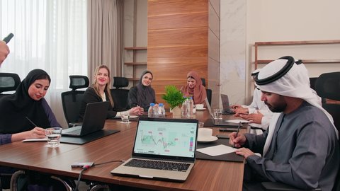 شرح وتوضيح العمليات والمشاريع والصفقات التجارية، التفاهم والتعاون بين الزملاء لتحقيق أهداف الشركة, نقاشات خاصة بالعمل, استخدام الحاسوب المحمول، اجتماع عمل مع موظفين عرب خليجيين اماراتيين مع رجال أعمال أجانب في غرفة الاجتماعات، ضمان فهم واضح وشامل للأعمال