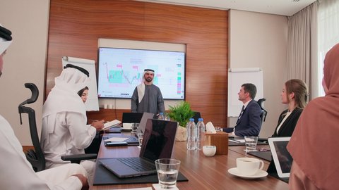 Completing tasks via computer, displaying statistical data on the screen, agreeing on business projects, a work meeting between Emirati Gulf Arab employees and foreign businessmen in the meeting room, the comfortable work environment in Emirati companies, using modern technological devices in offices.
