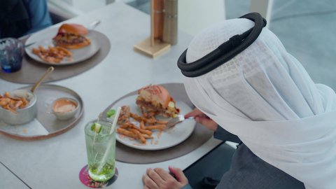 العمل عن بعد في احدى مطاعم الامارات العربية المتحدة, الاستمتاع بأجواء هادئه خارج مقر الشركة، تناول وجبة الغداء في الاستراحة، تصوير من الخلف مقرب لرجل اعمال عربي خليجي اماراتي يجلس على مائدة مليئة بالأطعمة، بناء علاقات اجتماعية ناجحة,