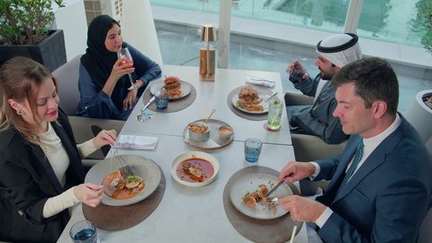 بناء علاقات اجتماعية ناجحة, مشاركة الأصدقاء الأوقات الممتعة, تبادل الأحاديث والاخبار, الاستمتاع بتناول الغداء في المطعم, مجموعة من الأصدقاء الاجانب والعرب خليجيين إماراتيين يجلسون على المائدة في إحدى المطاعم في الإمارات بإيماءات السعادة والسرور