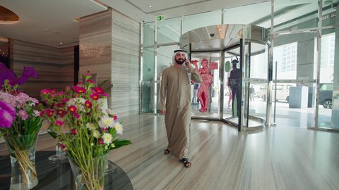Using modern technologies, making a phone call via mobile, enjoying shopping and browsing, acquiring necessities from malls, an Arab Gulf Emirati man wearing a kandura and ghutrah is wandering in the mall.