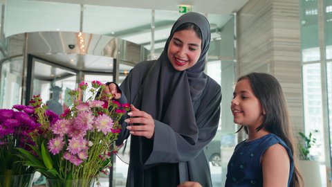 The concept of a happy family, feelings of motherhood and care, acquiring necessities from malls, an Arab Gulf Emirati woman placing a flower in her daughter's hair with expressions of love, spending enjoyable family time, benefiting from offers, discounts, and seasonal sales.
