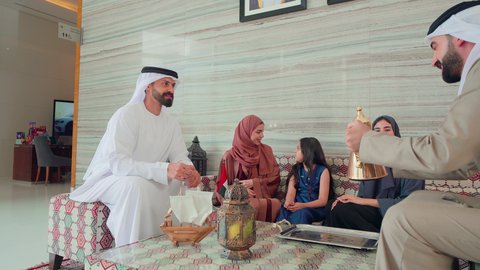 Spending happy times with family, an Emirati Gulf Arab man wearing a kandura and ghutra pouring coffee from a copper coffee pot, serving Arabic coffee, the concept of hospitality and reception, gestures of cheerfulness and welcoming guests