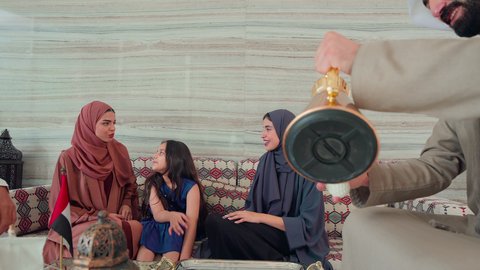 An Emirati Gulf Arab man wearing a kandura and ghutra pouring coffee from a copper coffee pot, enjoying hot drinks, smiling gestures and welcoming guests, the concept of hospitality and reception, serving Arabic coffee, spending happy times with family